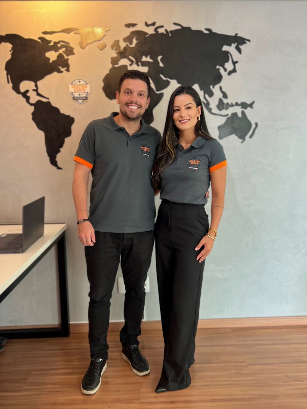 Os fundadores da Destino Certo Intercâmbios, de camisa da agência, em frente ao mapa-múndi do escritório