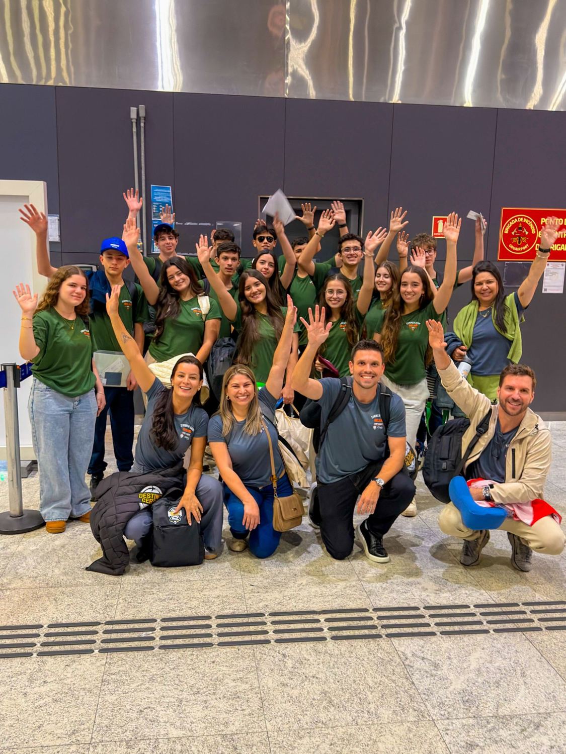 Grupo do Summer Camp acenando no aeroporto, com camisetas verdes